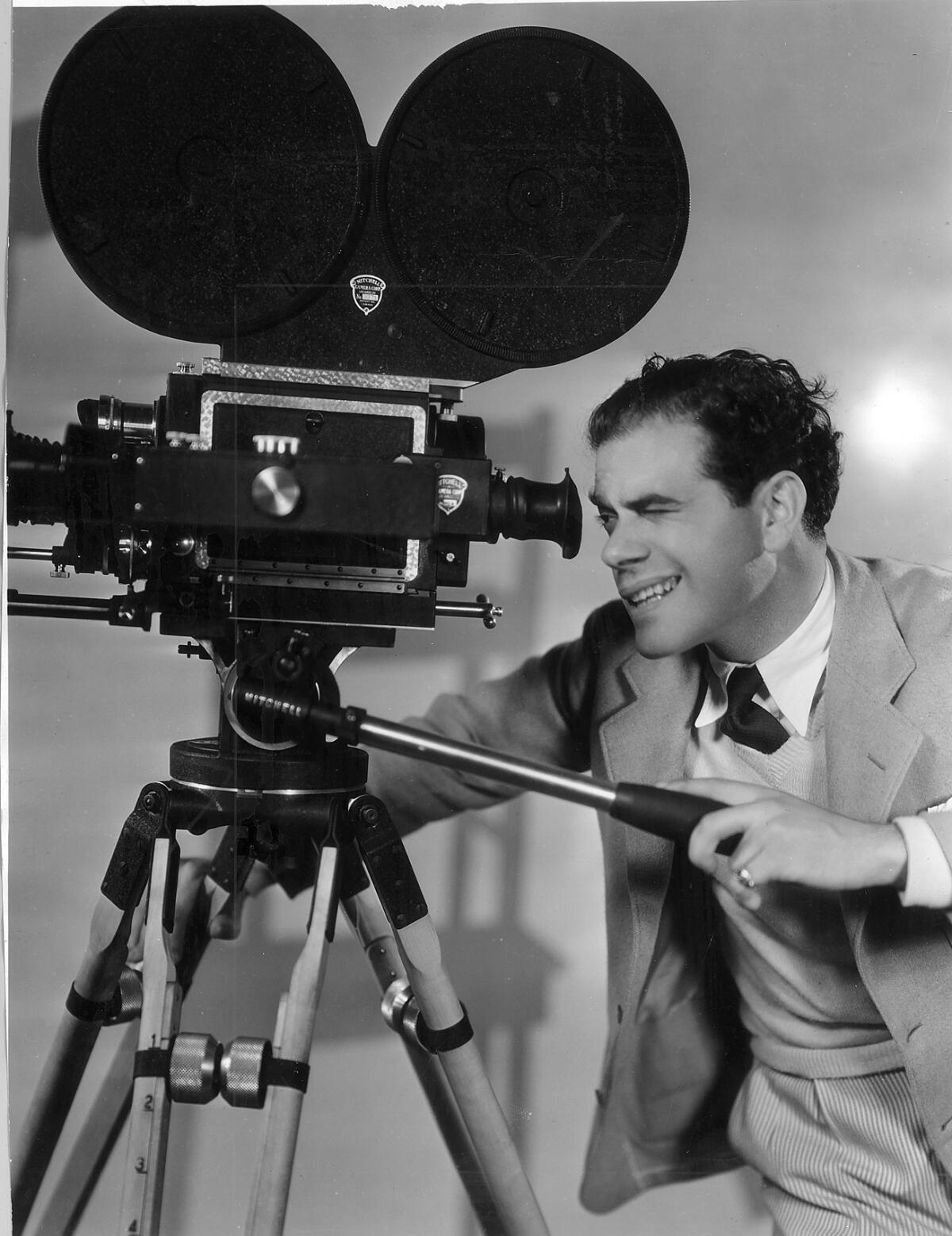 Director Frank Capra looks through the eyepiece of a movie camera on a tripod.