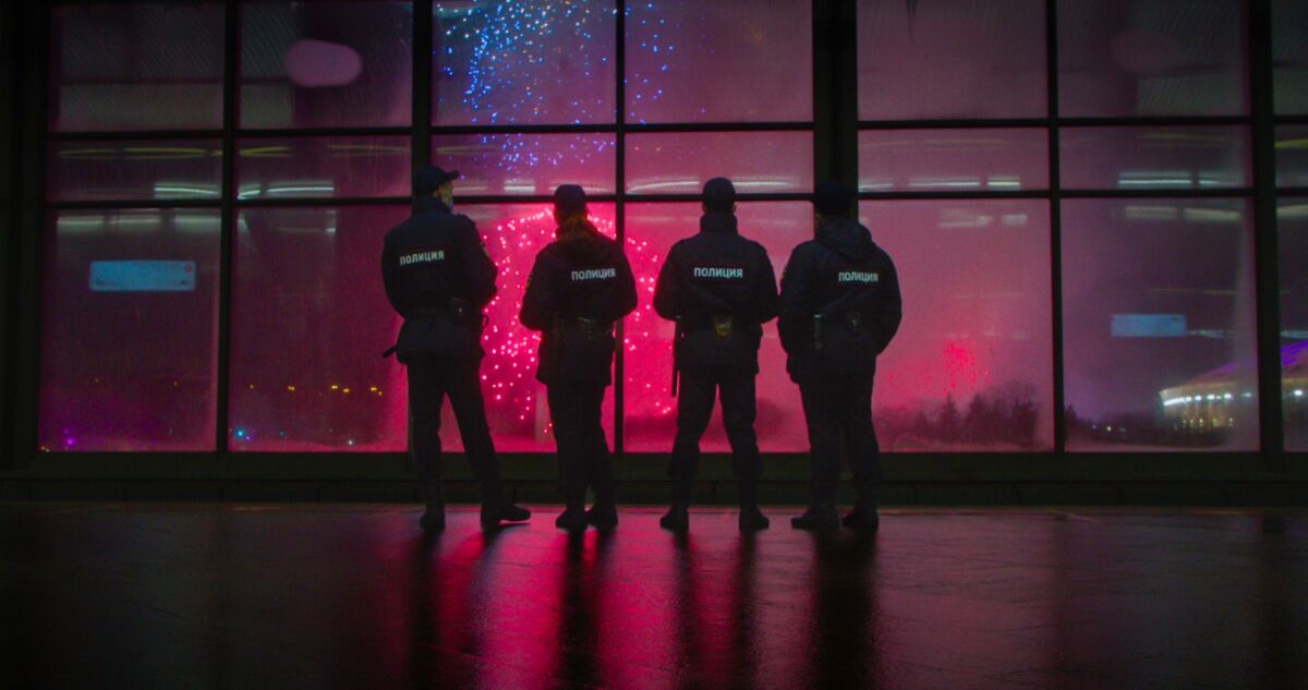 Four figures are silhouetted against floor-to-ceiling windows showing fireworks.
