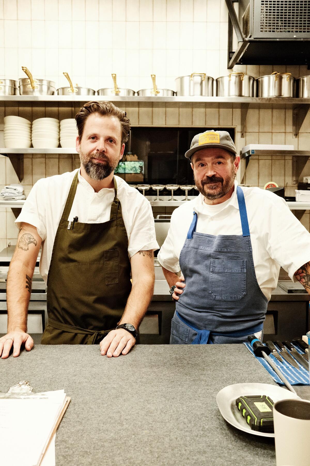 Two men in aprons stand behind a counter, looking at the camera, in a kitchen.