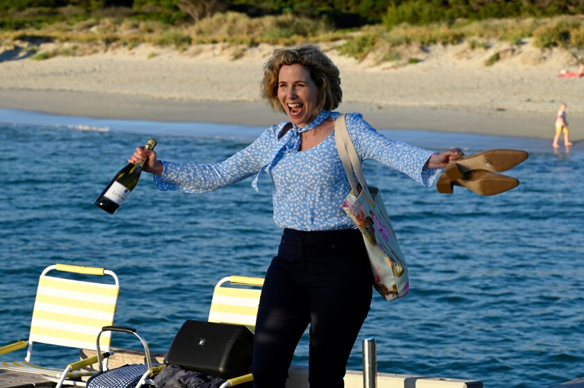 A smiling woman holding a wine bottle and her shoes at the seaside.