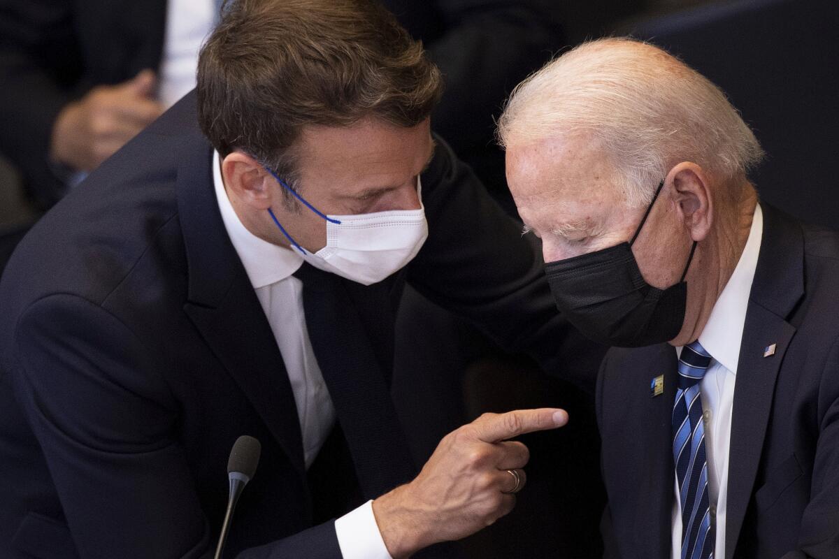 A masked man in dark suit and tie leans down and points a finger at another  man, right, also masked and in suit and tie
