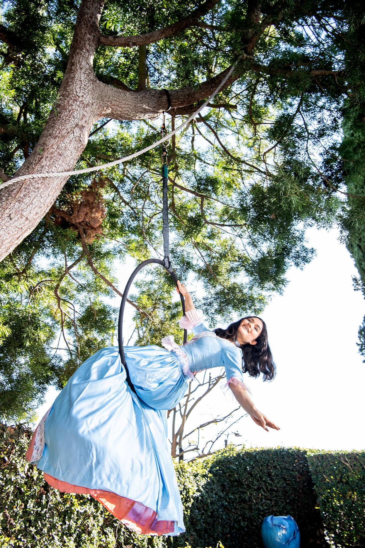 Rose, in a baby blue gown, sits on an aerial hoop, leaning back and hanging on with one hand.