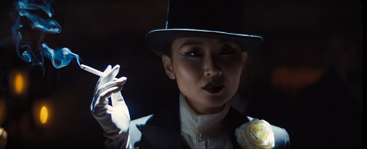 A woman, her face in shadow under the brim of her hand, holds a smoking cigarette in her white-gloved hand.