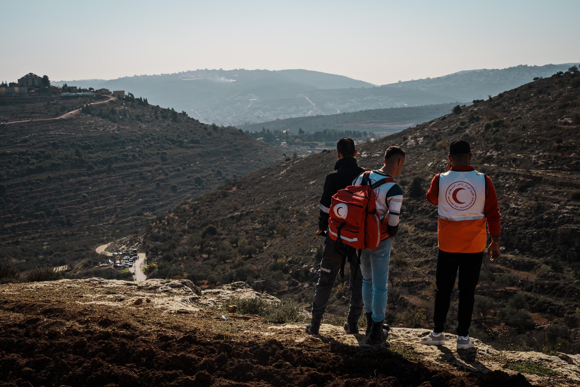 Three people, two with Red Crescent logos, stand on one of several hills and look out at the area below