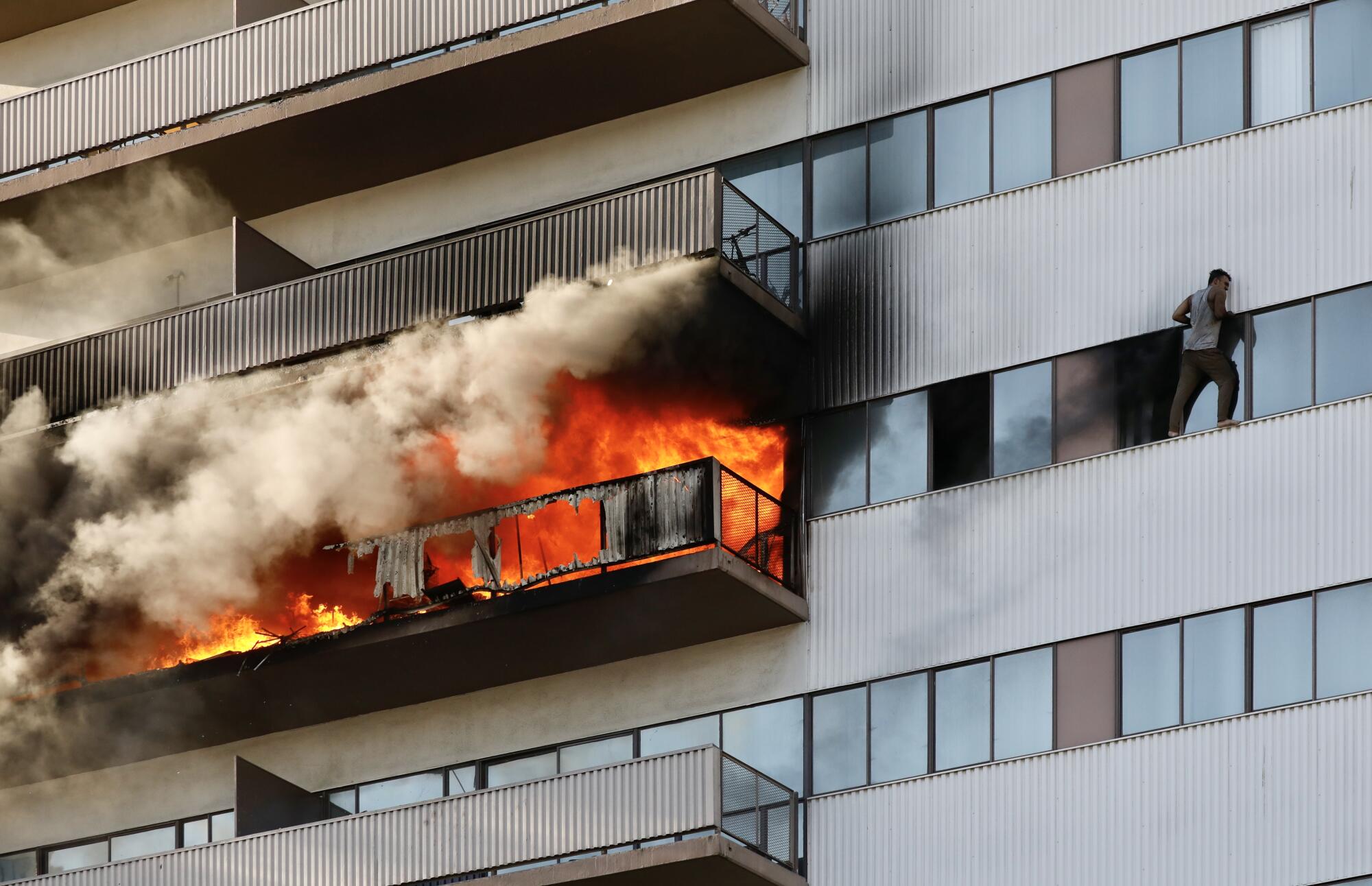 A resident, arms blackened, stands on the ledge of a window several stories up. Fire rages on a balcony near him.