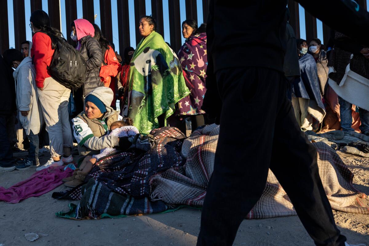 People standing and sitting bundled up alongside a tall border fence
