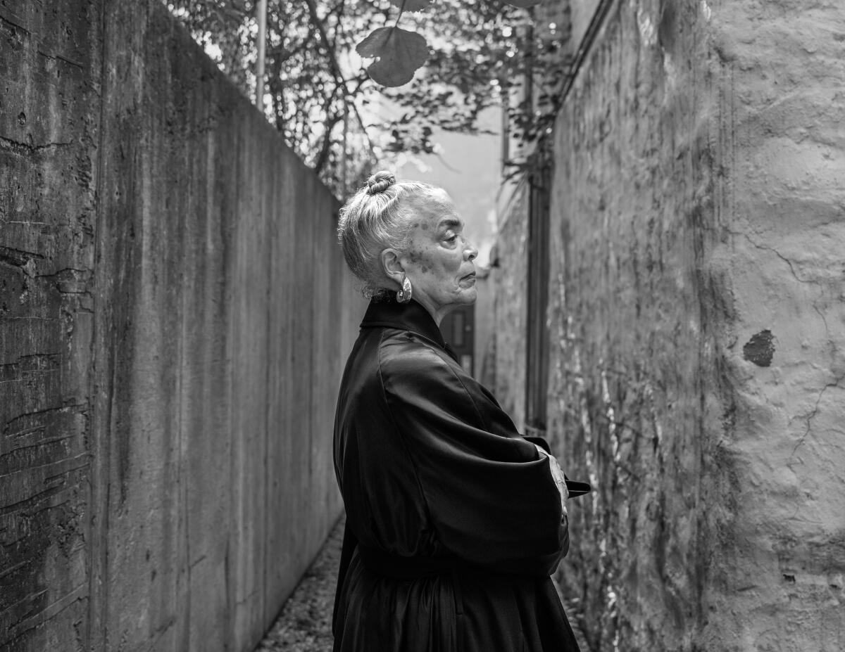 Maren Hassinger, is seen wearing a dark overcoat in profile between two concrete walls in a black and white photo.