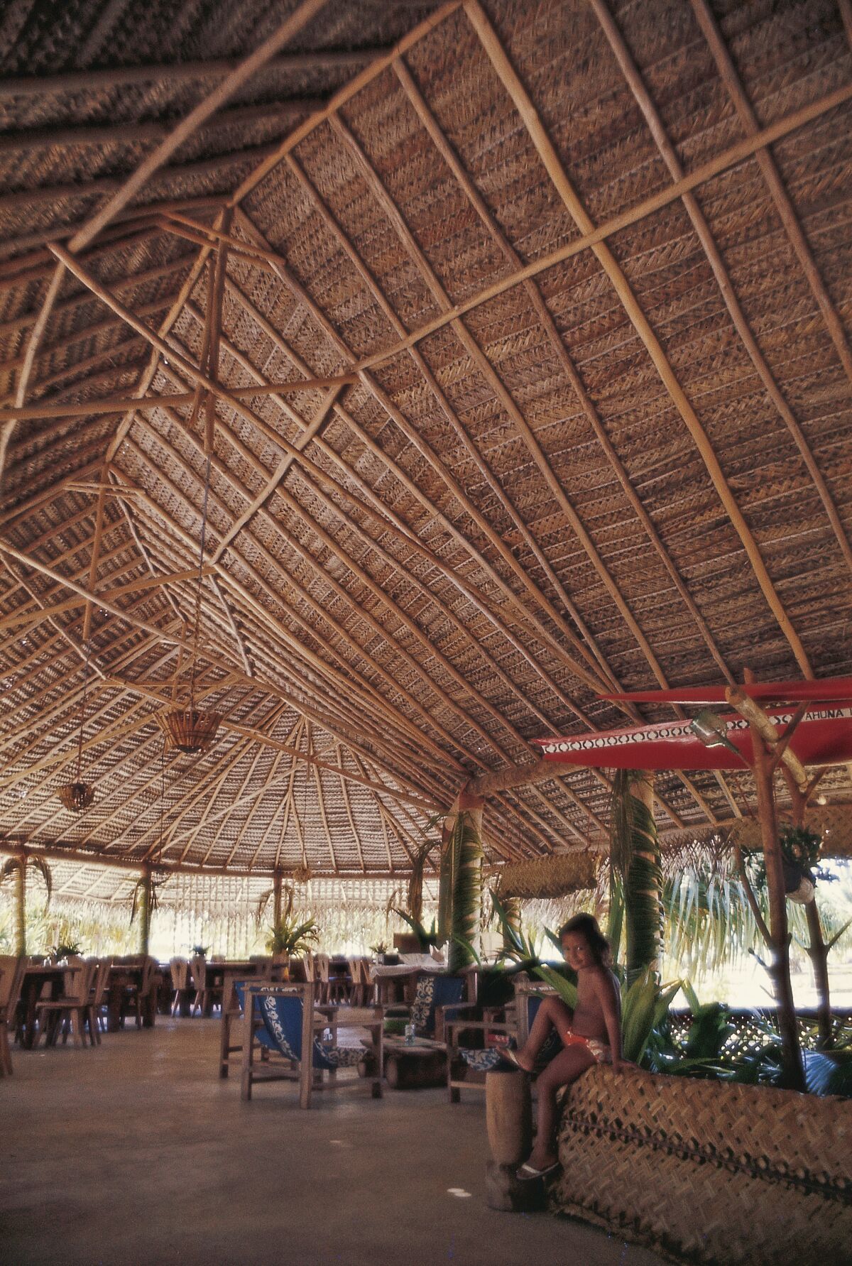 A child sits in a large open-air pavilion inspired by Polynesian architecture, made from bamboo and woven palm fronds.