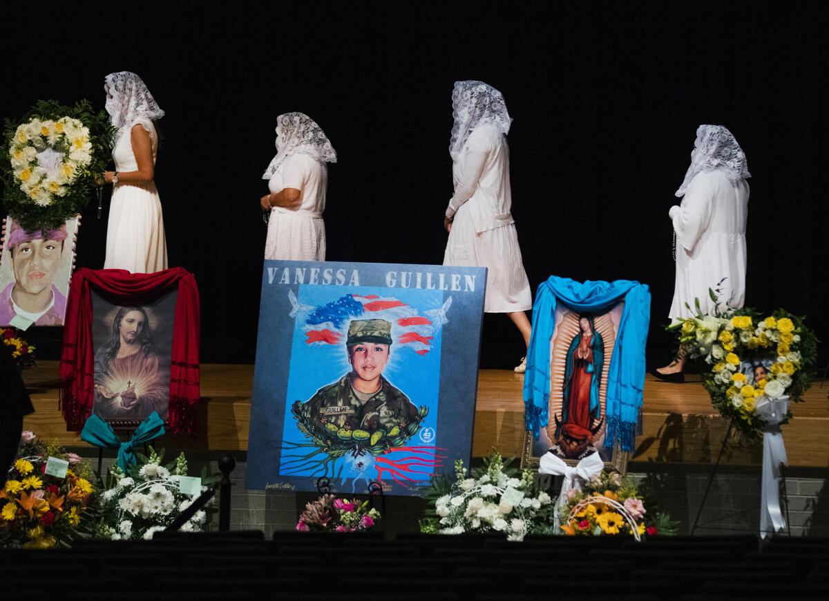 Four women in white with headscarves walk onstage  behind flowers and portraits of Jesus, Mary and Vanessa Guillén in uniform