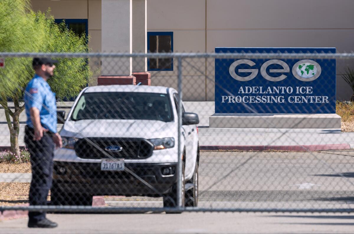 A person stands behind a gate outside a building with a sign that reads "GEO Adelanto ICE Processing Center."