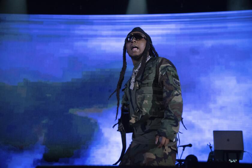 A man with long dark braids performs onstage with a blue background
