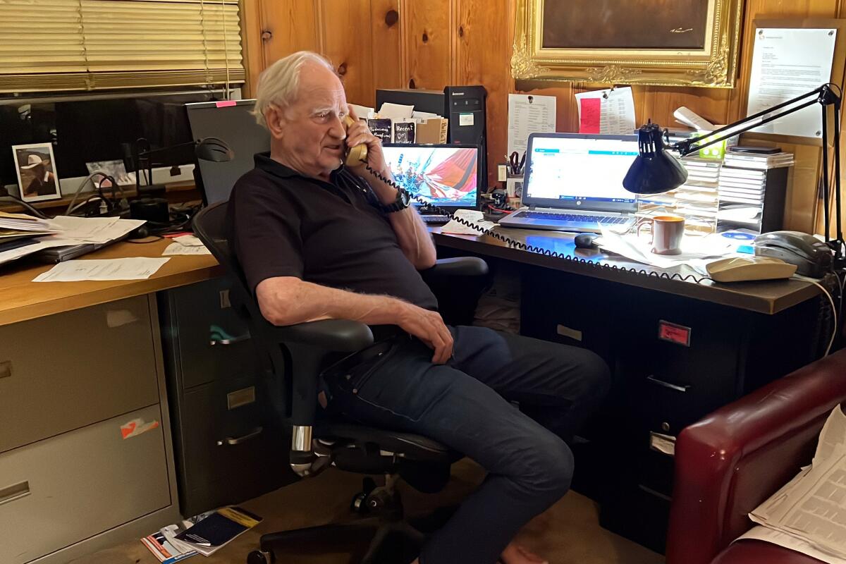 A man in dark polo shirt and pants seated at a desk speaks on a landline phone