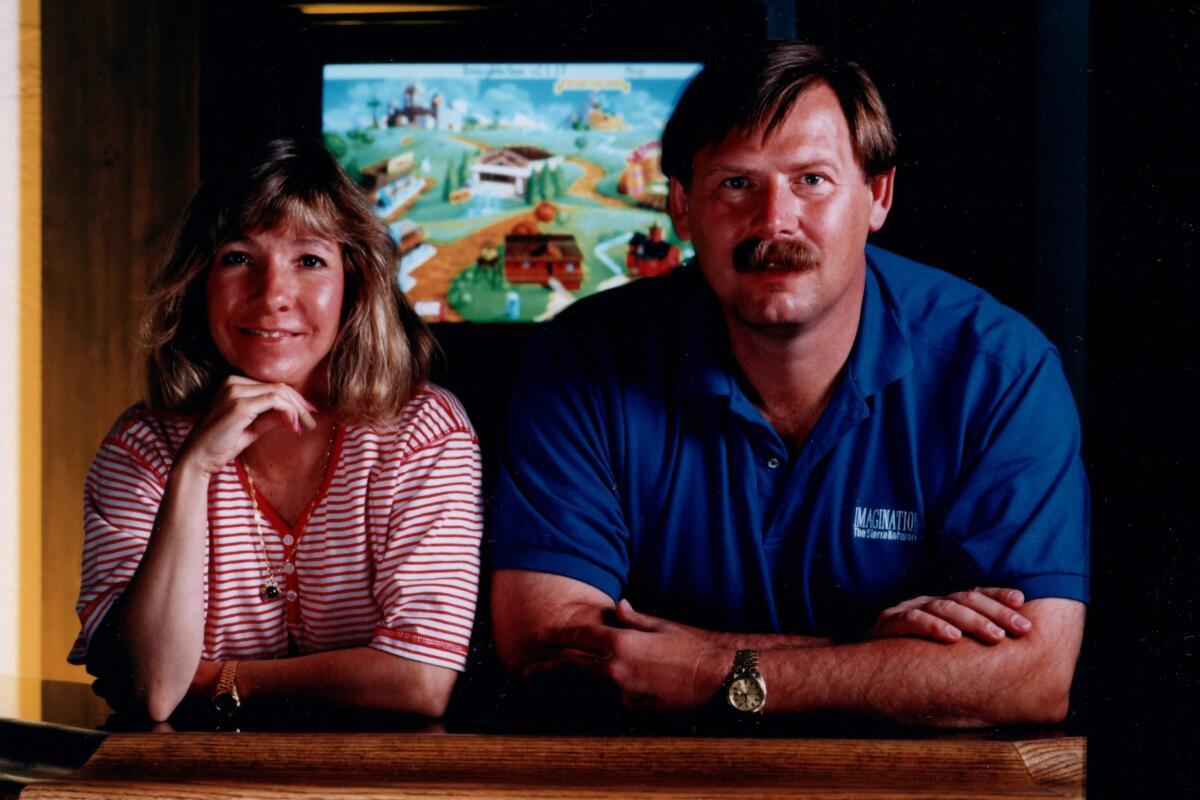 A woman and a man lean forward, elbows on a counter, with a video game on a monitor behind them.