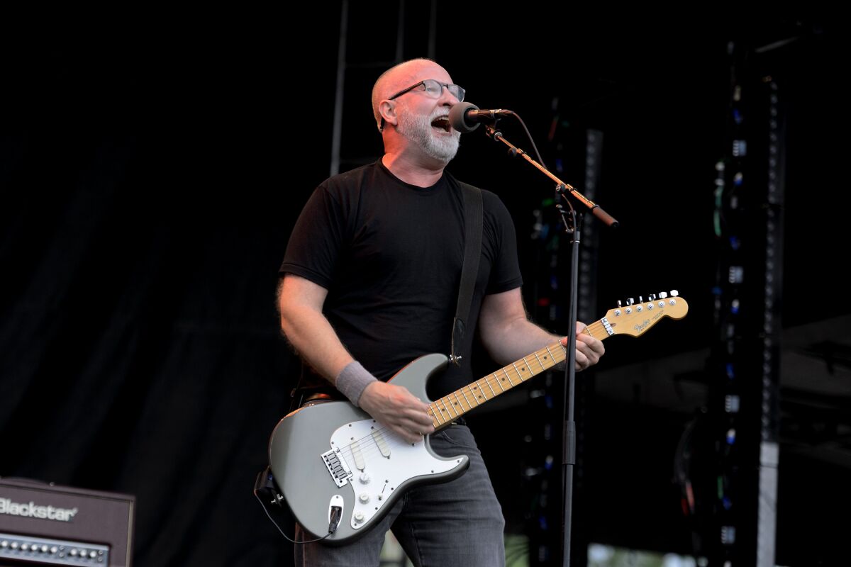 A man with a gray beard and glasses and wearing a black T-shirt plays electric guitar and sings into a microphone on stage. 