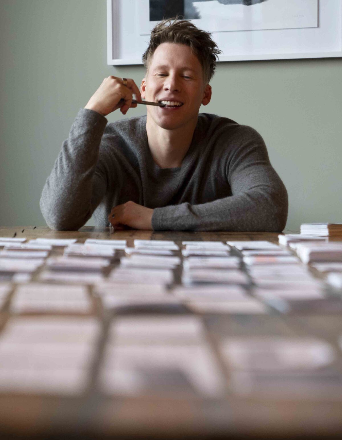A man with a pen in his mouth sits at a table covered in index cards