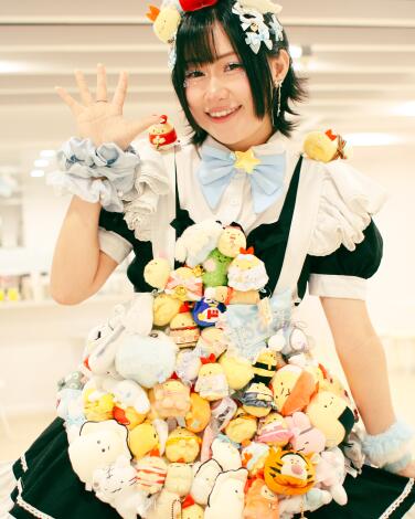 A woman dressed in a maid outfit covered in plush stuffed animals holds her fingers up in a peace sign.