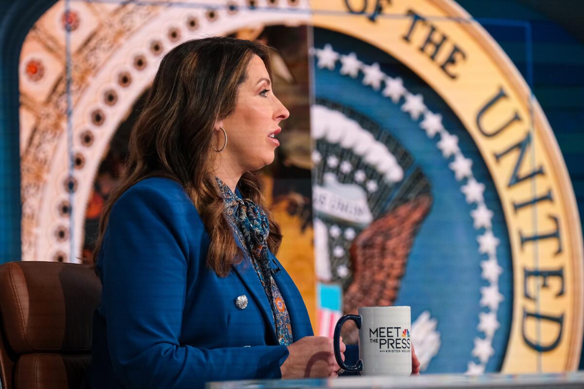 A woman in front of the presidential seal holding a mug that says 'Meet the Press'