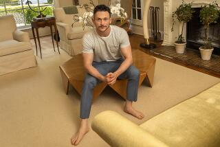 Actor Jonathan Tucker sits on a wooden coffee table near a fireplace, sofa and chairs in a beige-toned living room