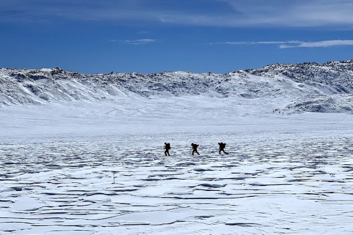 Three small figures walk on skis across a snow-covered landscape with small mountains on the horizon.