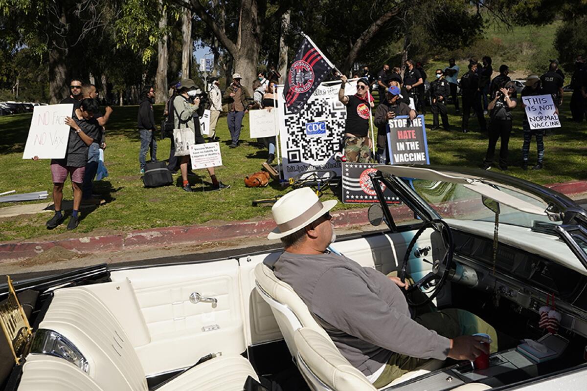 A car passes a group holding flags and signs that read "Stop the Hate in the Vaccine" and "This is Not a Normal Vaccine"