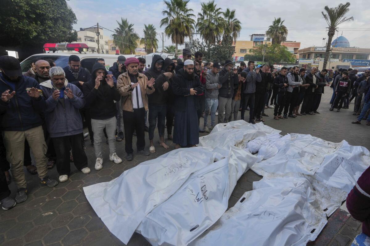 Mourners stand in a line, holding up their palms in prayer, facing bodies covered in white sheets on the ground 