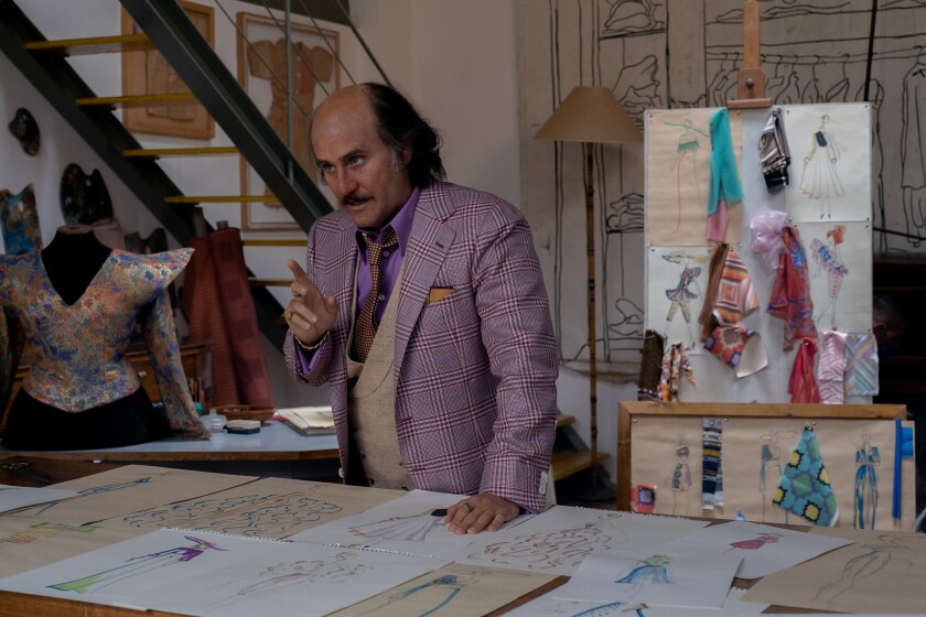 A balding man in a purple plaid suit stands in front of a table full of fashion sketches.