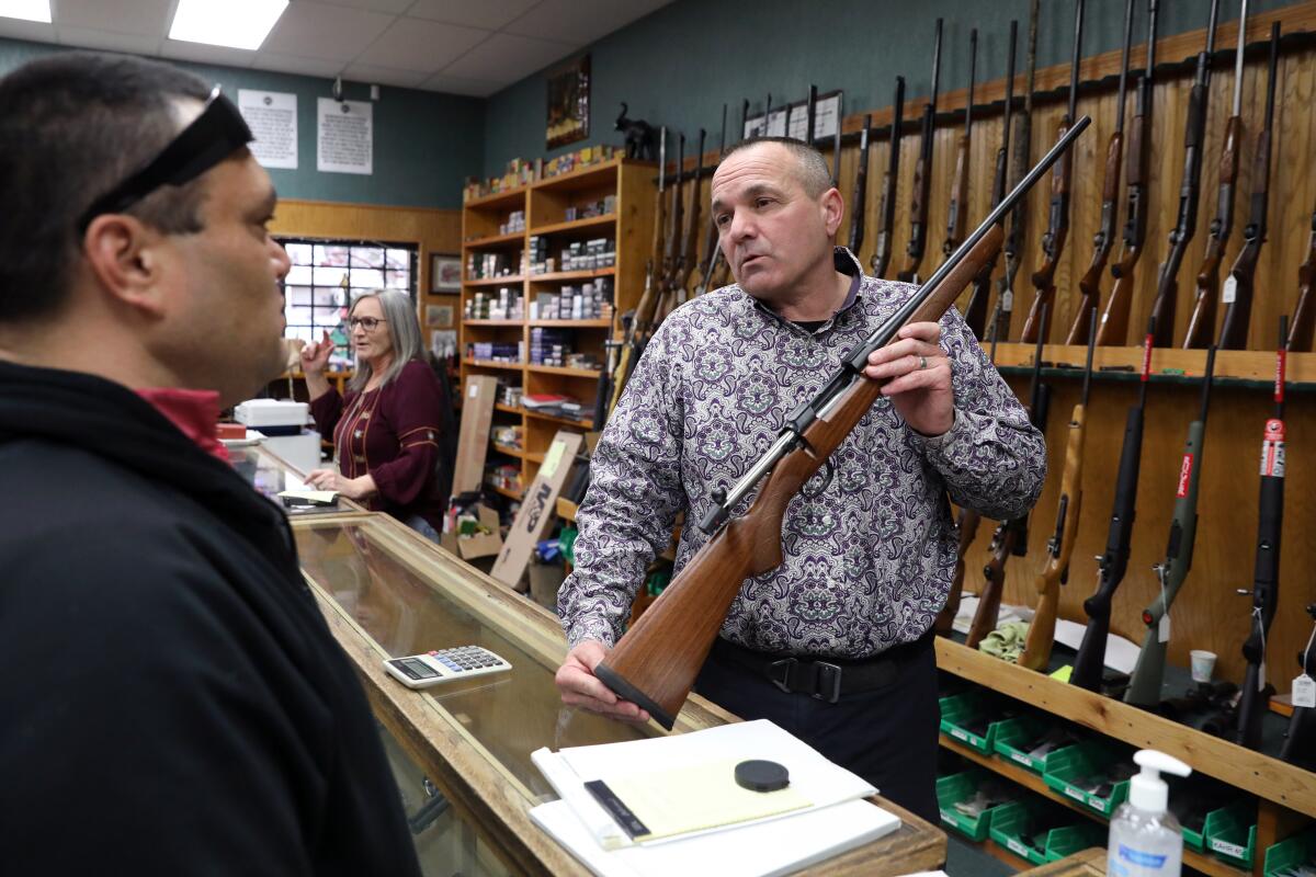 A man behind a counter holds a rifle up for a prospective customer.