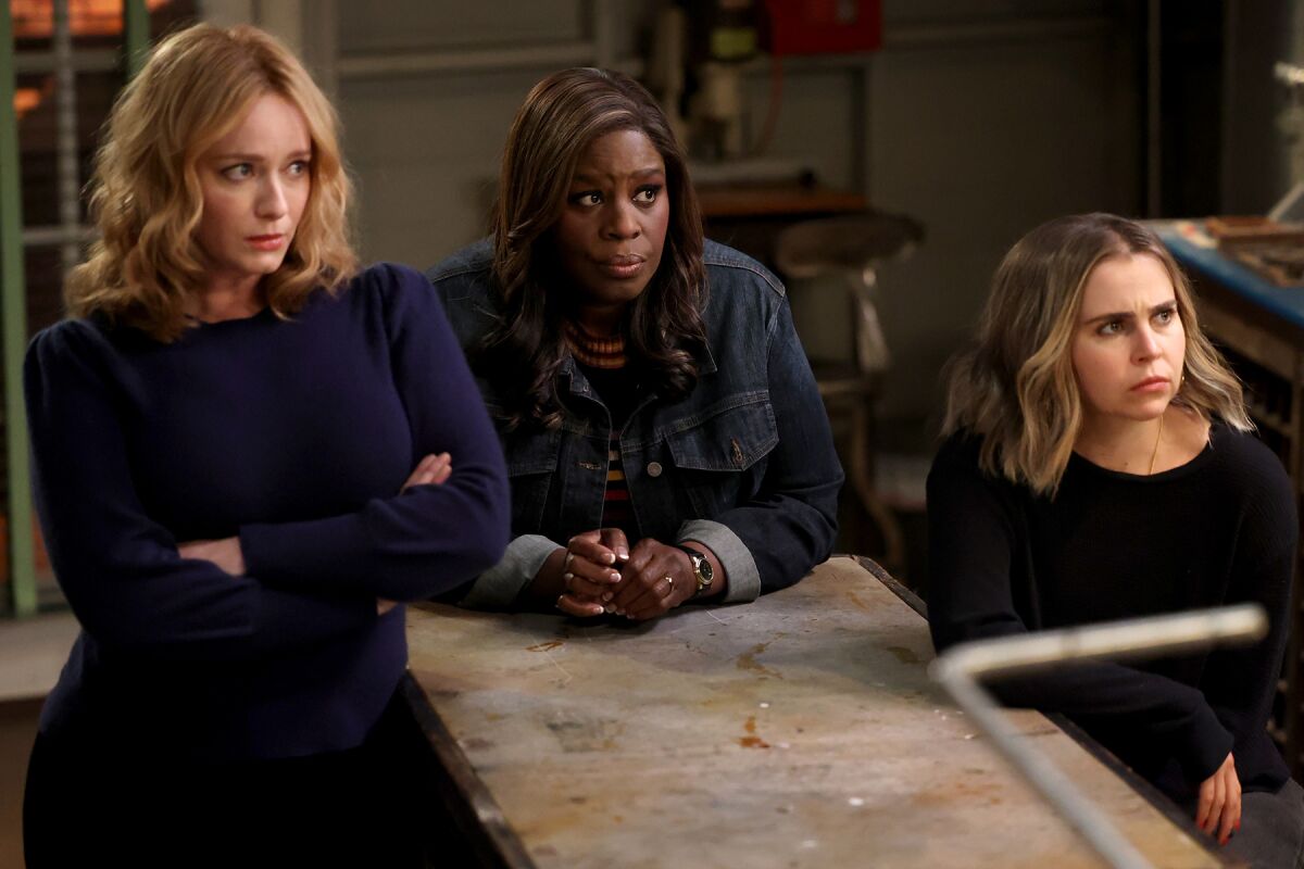Three women gather alongside a kitchen counter looking apprehensive.