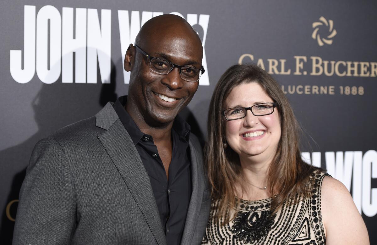 A bald man with glasses in a suit smiles and poses with a smiling woman in a patterned dress.