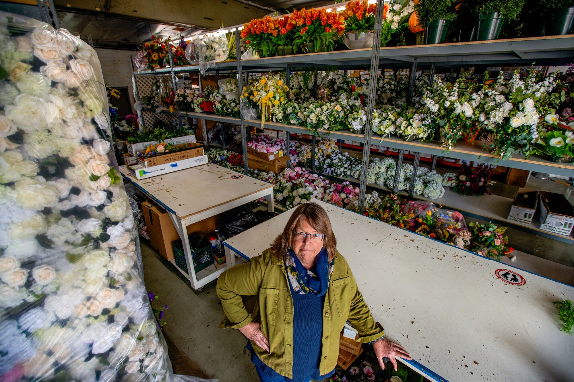A woman leans on a table next to silk flowers, inside a warehouse.