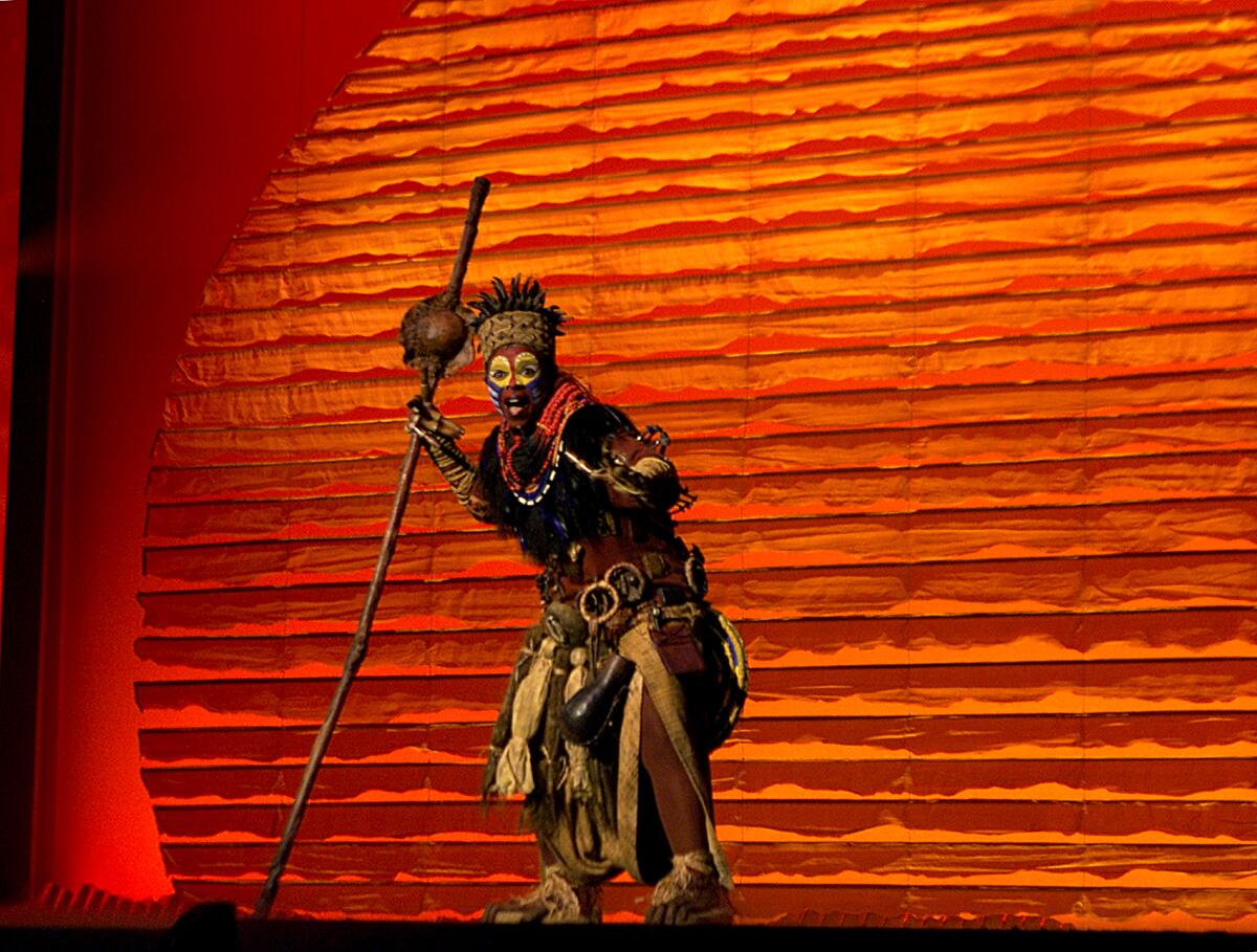 A performer holding a long staff and wearing heavy makeup stands on stage against a red background.
