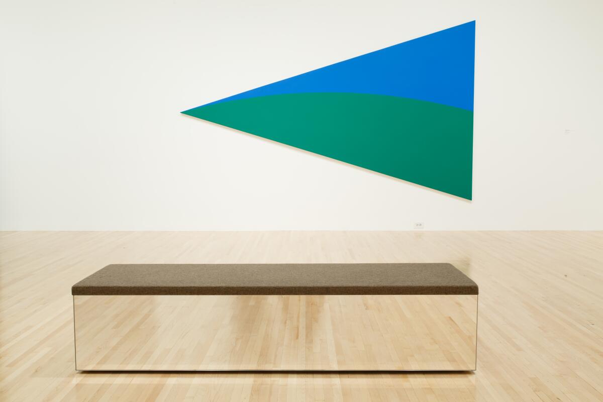 A mirrored bench topped with carpet in a wood floor gallery where a blue/green painting of a triangle hangs.