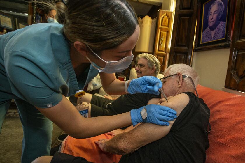 LOS ANGELES, CA-OCTOBER 7, 2022: Licensed Vocational Nurse Angela Tapia administers a dose of the Moderna COVID-19 booster vaccine to Louis Salazar Sr.,90, at his home in Los Angeles. In background is Salazar's son, Louis Jr., 64, who received the booster shot first. The Los Angeles County Department of Public Health runs a program that provide COVID vaccines to people who are homebound. (Mel Melcon / Los Angeles Times)