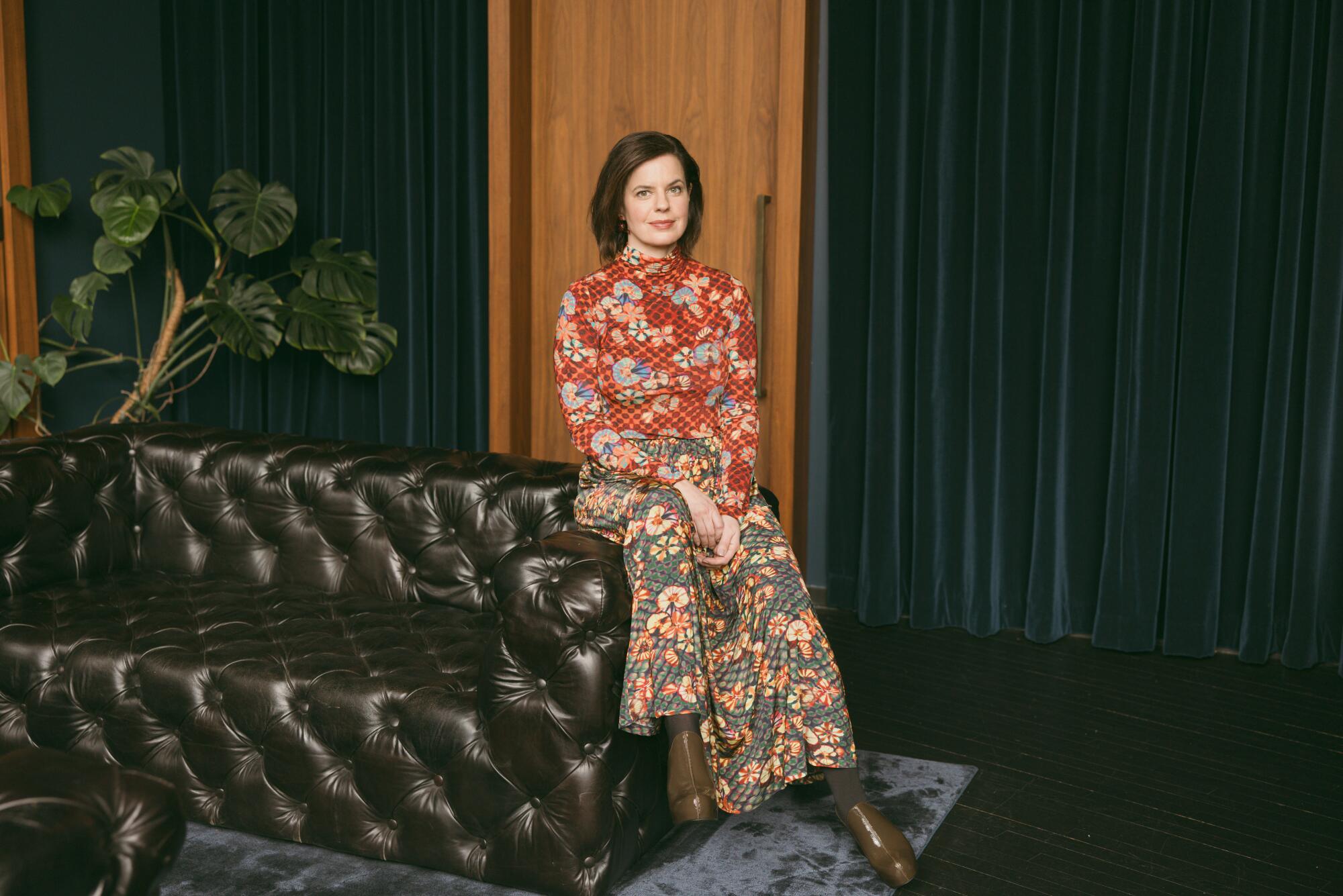 A woman in a floral top and long skirt is sitting on the edge of a brown leather sofa.