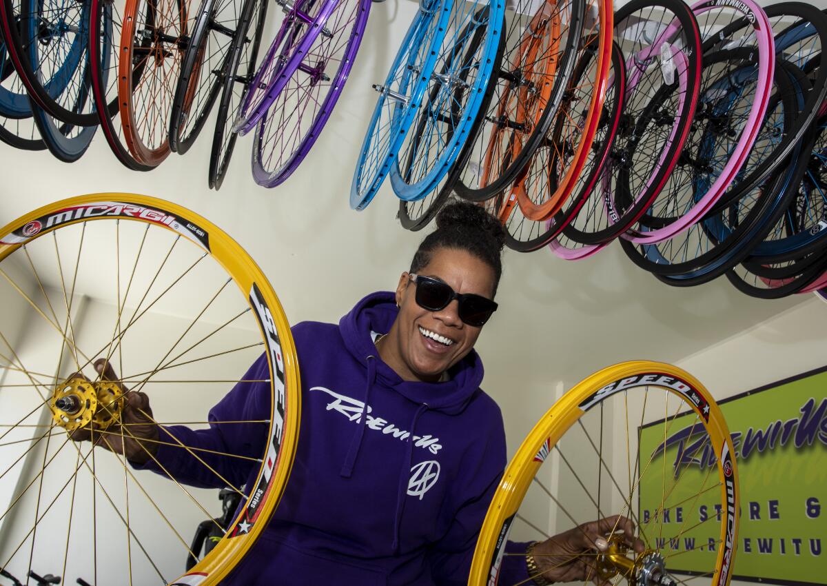 A woman smiles while holding two yellow bike wheels in front of a wall of multicolored wheels