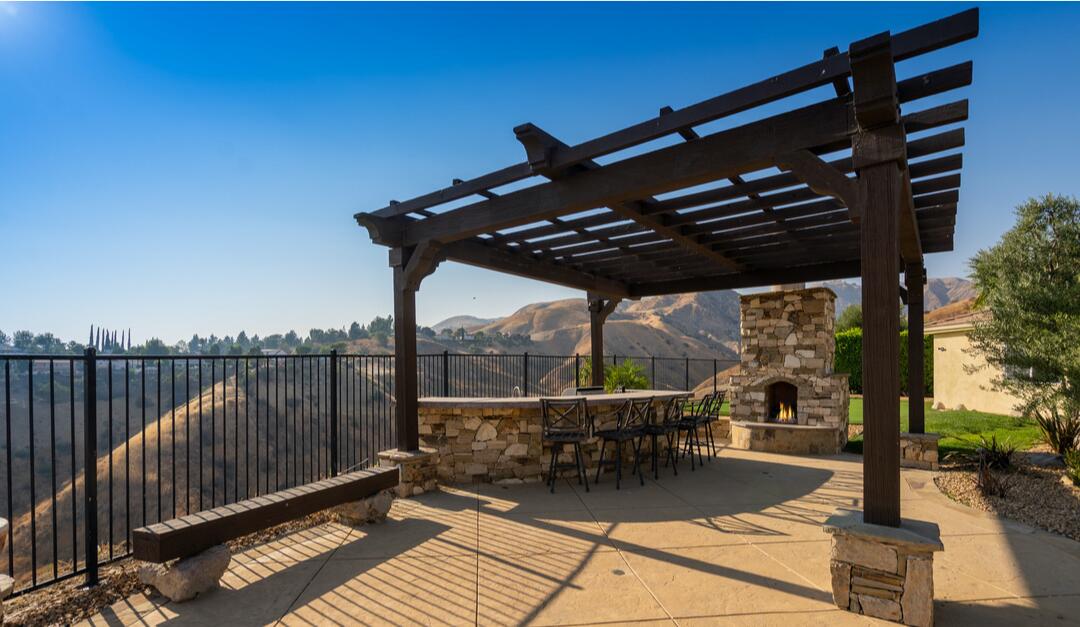 A pergola with a slatted roof covers a barbecue area and stone-covered bar alongside an outdoor fireplace.