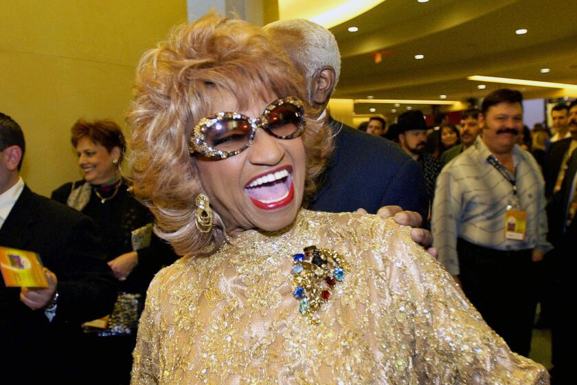 woman, celia cruz, smiling with mouth open, with curled blond hair, in large sunglasses and wearing gold, shimmering dress