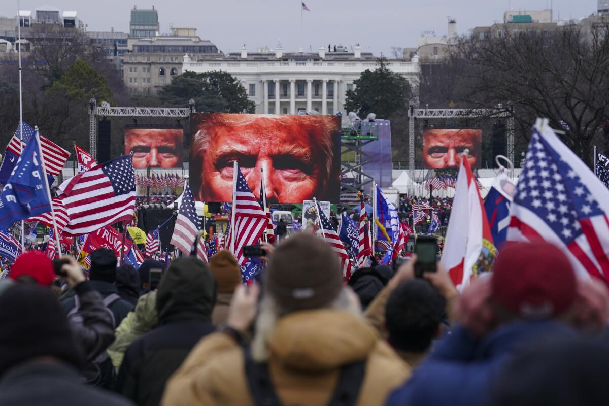 Trump supporters hold flags and banners while three big screens show the top half of Trump's face.