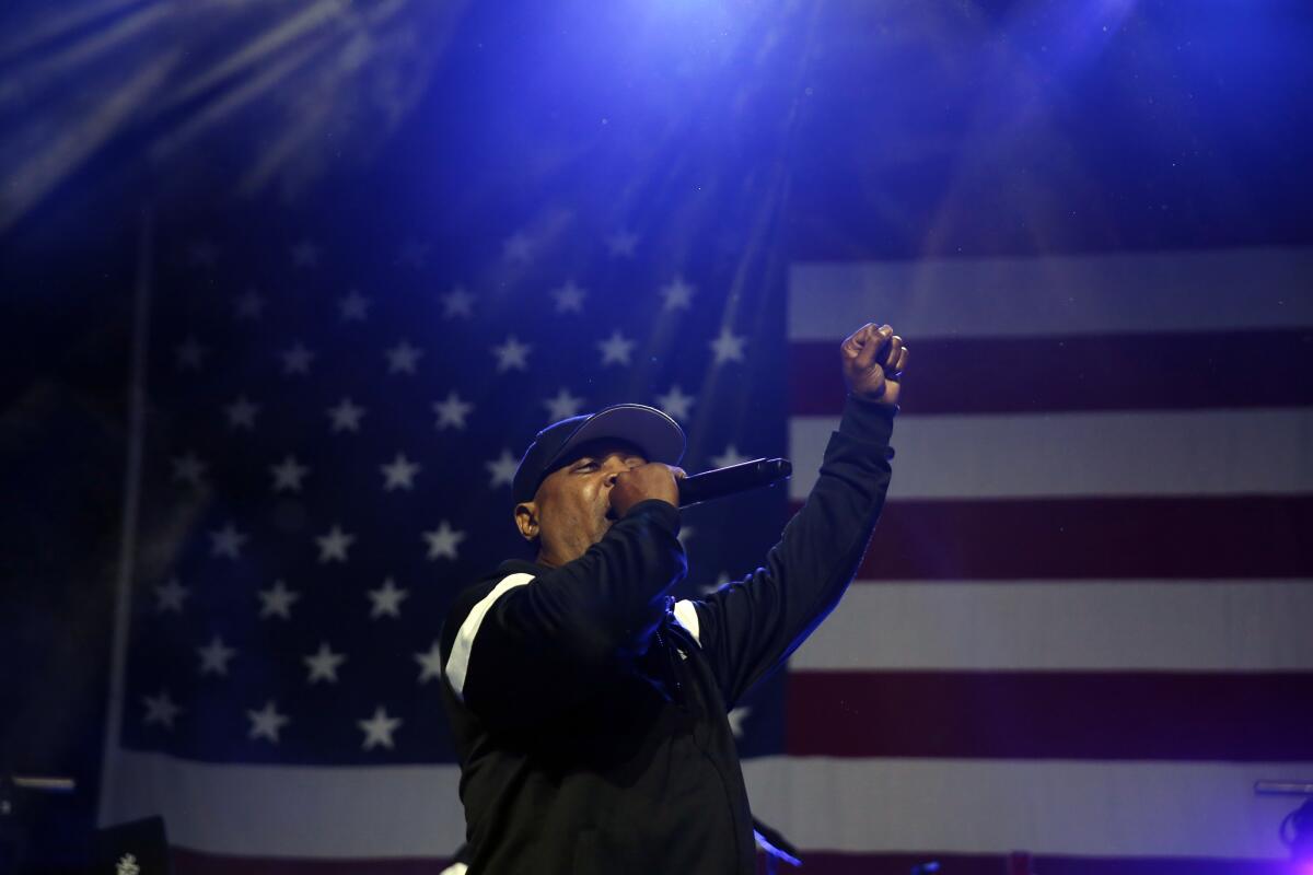 A man wearing a black tracksuit sings into microphone on stage with his fist raised in front of an American flag