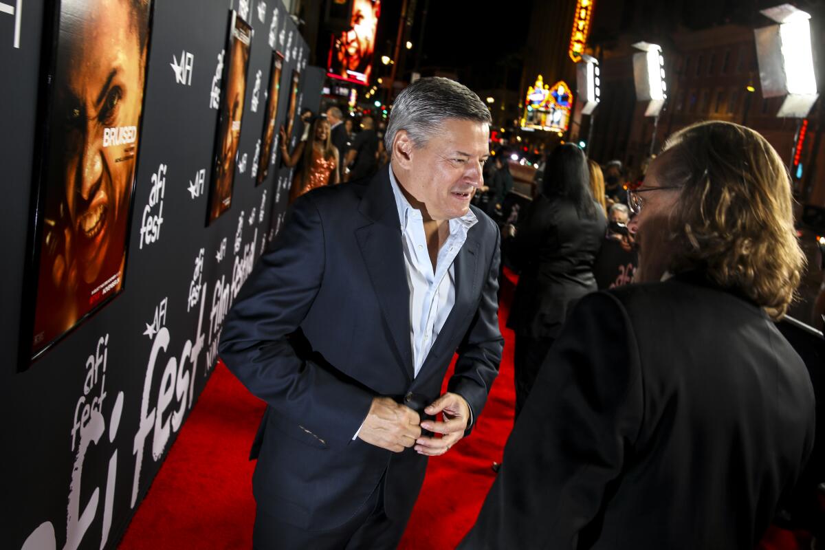 Two men in dark suits talk on a red carpet.