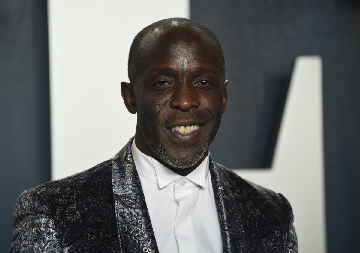 Michael K. Williams in a silver suit jacket and a dress shirt smiling in front of a grey background