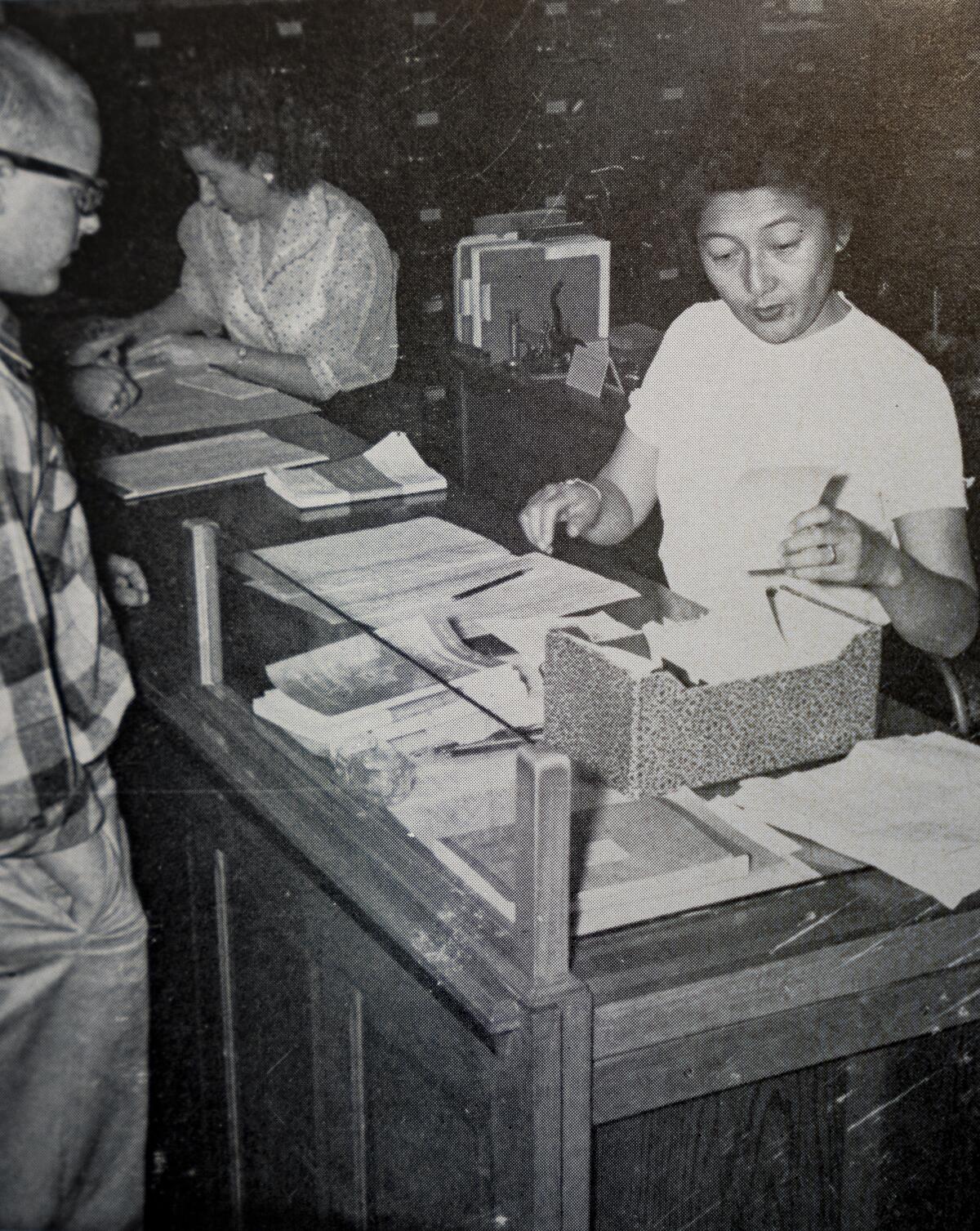 A woman sorts through documents on a table as a man waits. 