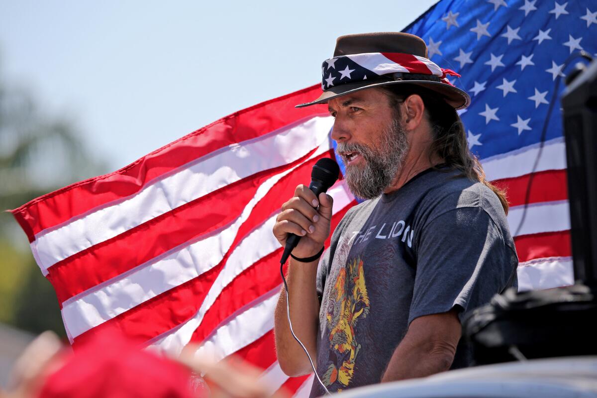 A man with a U.S. flag bandanna on his hat speaks into a microphone as another U.S. flag flies behind him