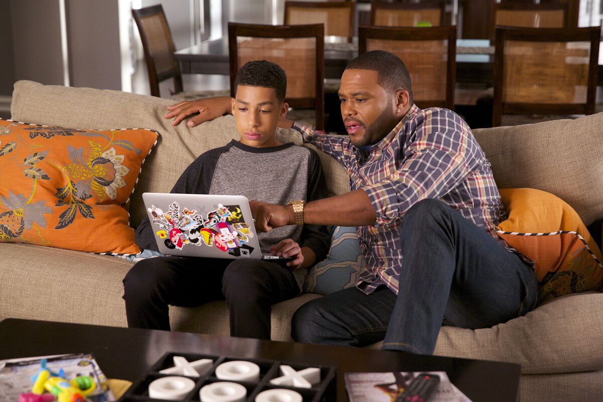 A man sitting on a couch, pointing at the laptop screen of his teenage son sitting next to him.