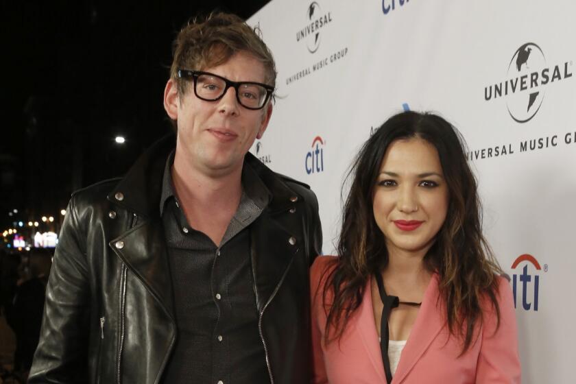 A blond man wearing glasses and a black leather jacket posing with a brunette woman wearing a pink blazer