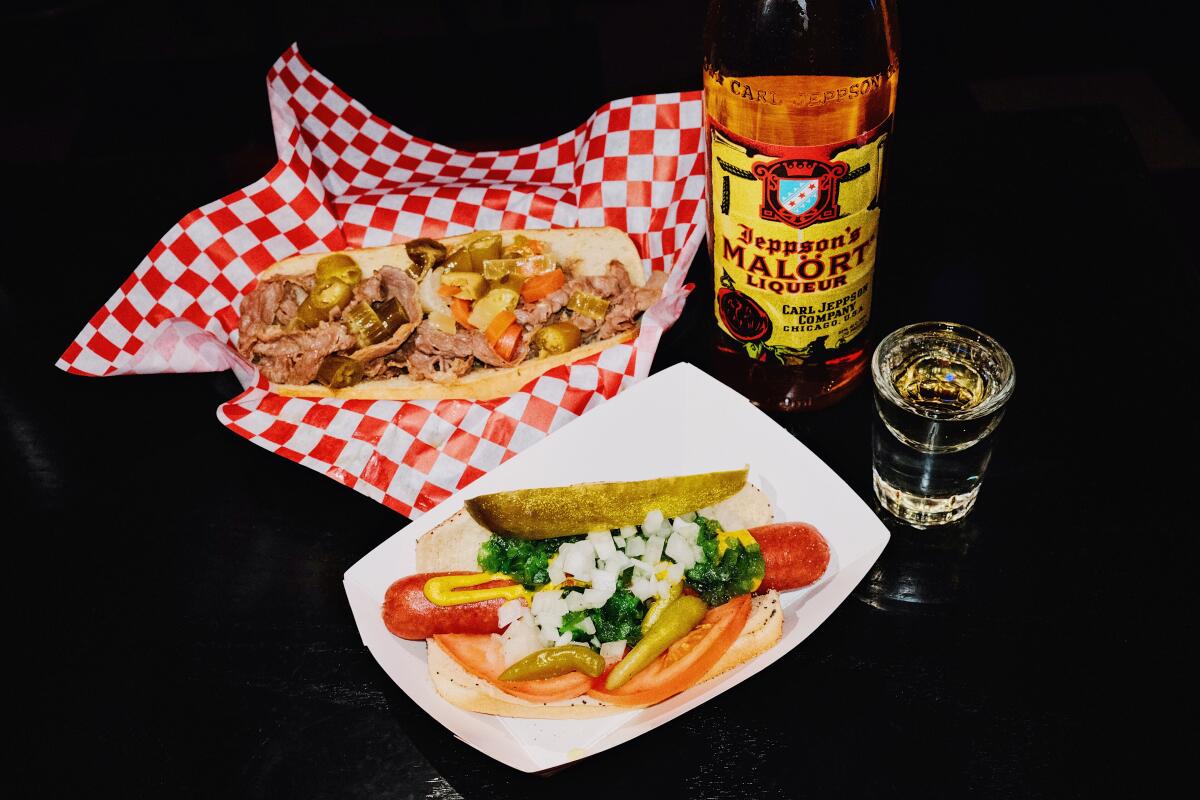 An Italian beef sandwich with a Chicago dog and a bottle and shot of Jeppson's Mal?rt on a black table.