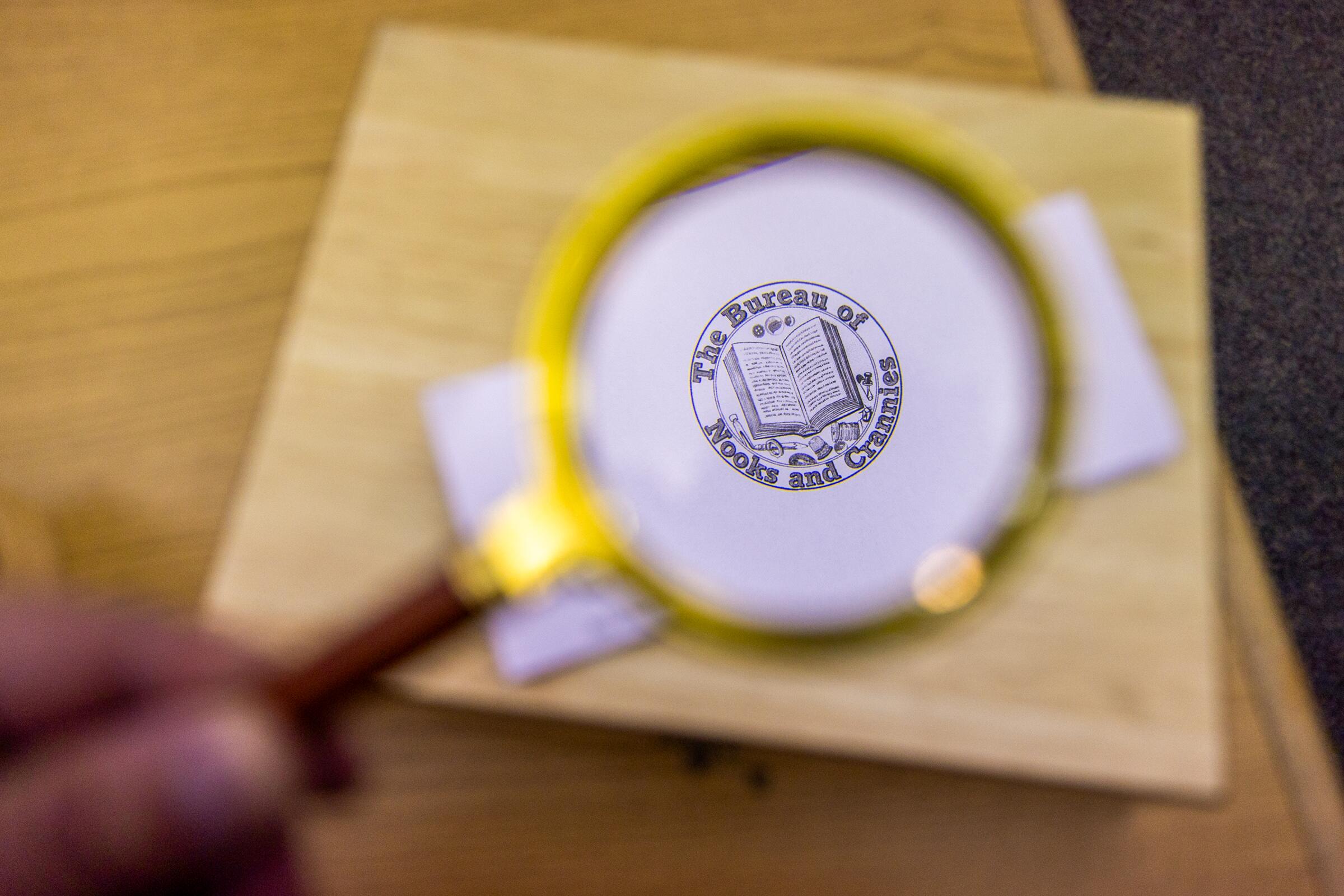 A hand holding up a magnifying glass, which highlights a stamp labeled "The Bureau of Nooks and Crannies"