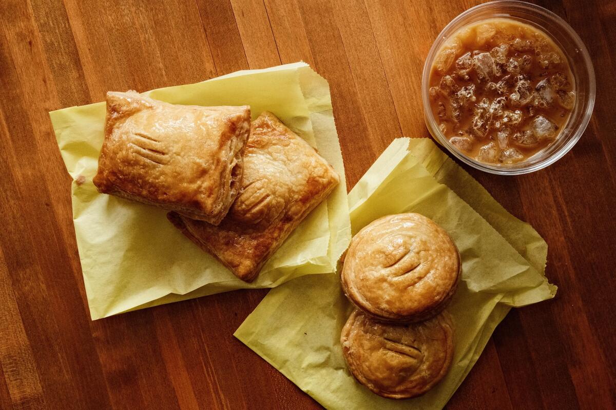 Four sweet and savory pastelitos atop yellow bags, with an iced cafe con leche, on a tabletop at Cafe Tropical