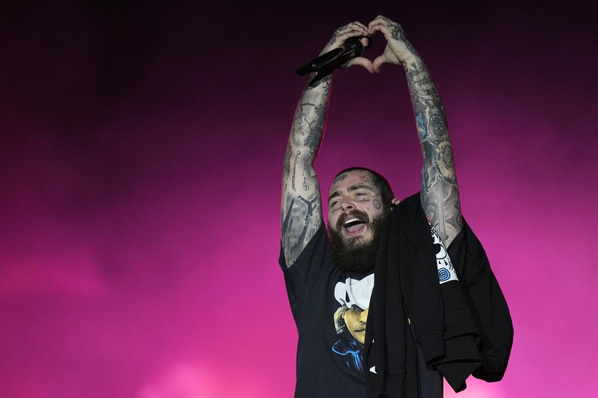 Post Malone in a black T-shirt forming a heart with his hands above his head onstage