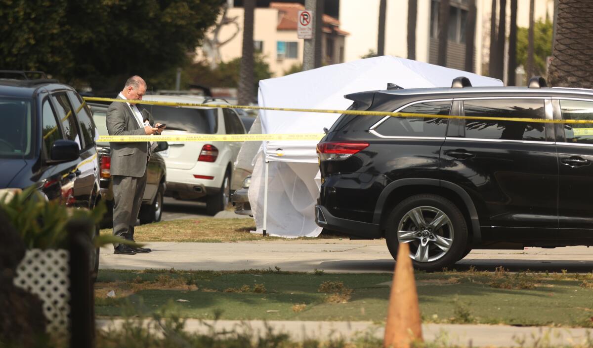 A man in a suit standing behind yellow crime scene tape near shell casings, a white tent and several parked vehicles